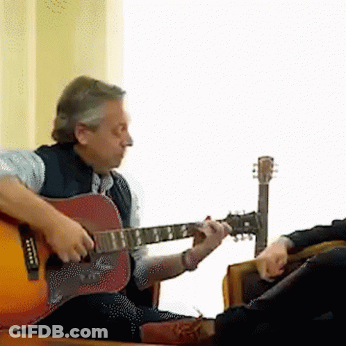 A Man Is Playing An Acoustic Guitar While Another Man Sits In A Chair GIF