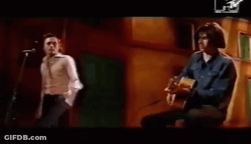 A Man Is Playing An Acoustic Guitar While Another Man Stands Behind Him . GIF