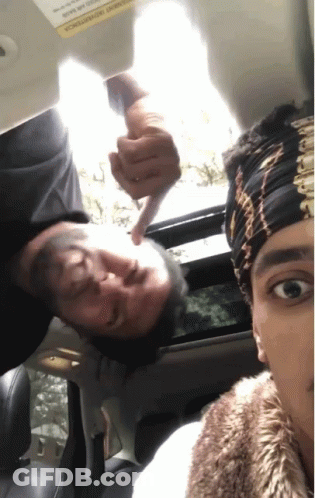 A Man Is Pointing At Another Man 'S Face In A Car With A Warning Sign On The Visor GIF