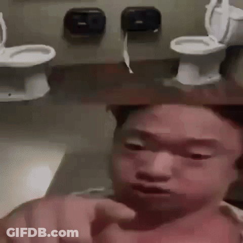 A Man Is Pointing At The Camera In A Bathroom With Toilets Behind Him . GIF