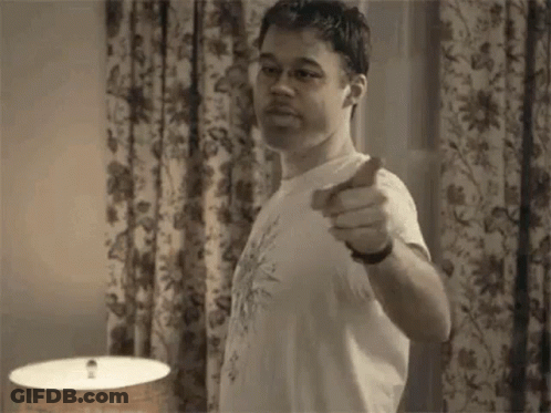 A Man Is Pointing At The Camera While Standing In Front Of A Lamp In A Room . GIF