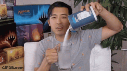A Man Is Pouring Milk Into A Glass In Front Of Boxes Of Magic The Gathering Cards GIF