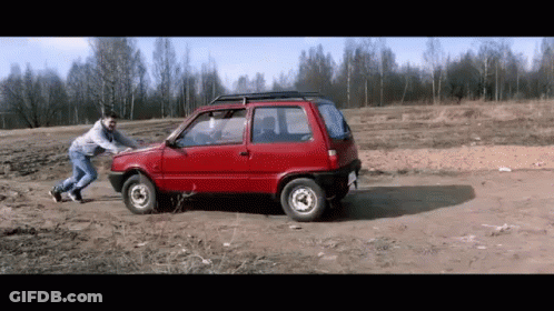 A Man Is Pushing A Small Red Car In A Dirt Field GIF