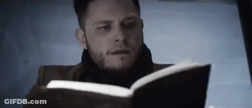 A Man Is Reading A Book With A Scarf Around His Neck . GIF
