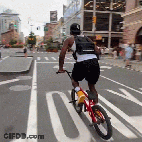 A Man Is Riding A Bike Down A Street With The Letter S On His Back GIF