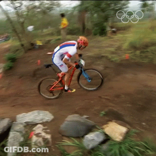 A Man Is Riding A Bike On A Dirt Trail With The Olympic Rings In The Background GIF