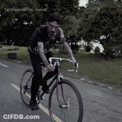 A Man Is Riding A Bike On A Road With La Guarimba Film Festival Written On The Bottom Right GIF