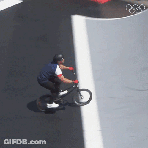 A Man Is Riding A Bike On A Road With The Olympic Rings In The Background GIF