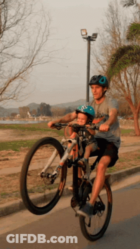 A Man Is Riding A Bike With A Child In A Helmet On The Back GIF