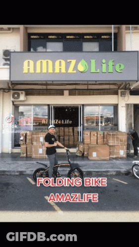 A Man Is Riding A Folding Bike In Front Of A Building That Says Amazlife GIF