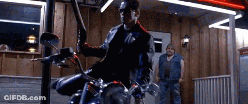 A Man Is Riding A Motorcycle In Front Of A Building While Another Man Stands Behind Him . GIF