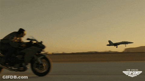 A Man Is Riding A Motorcycle In Front Of An Airplane That Says Top Gun Maverick GIF