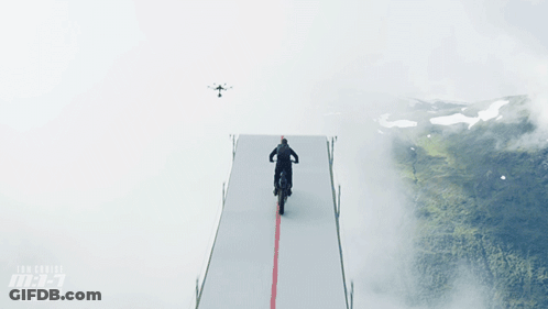 A Man Is Riding A Motorcycle On A Ramp With A Drone Flying Overhead GIF