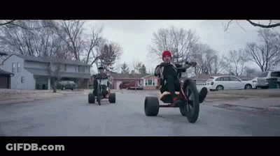 A Man Is Riding A Tricycle Down A Street Next To A Man Riding A Bike . GIF