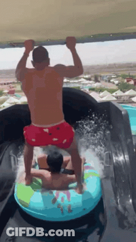 A Man Is Riding A Tube Down A Water Slide While Another Man Stands On Top Of It . GIF