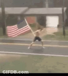 A Man Is Running Down The Street With An American Flag In His Hand . GIF