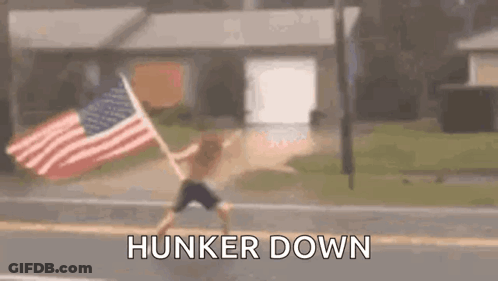 A Man Is Running Down The Street With An American Flag In His Hand . GIF