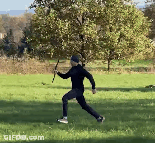 A Man Is Running In A Field With A Stick In His Hand . GIF