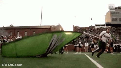 A Man Is Running On A Football Field With A Large Green Flag That Says ' M ' On It GIF