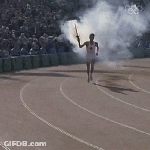 A Man Is Running With A Torch On A Track With The Olympic Rings In The Background GIF