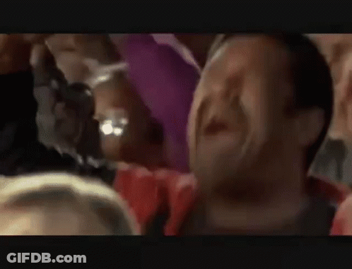 A Man Is Screaming In A Crowd Of People While Holding His Arm In The Air . GIF