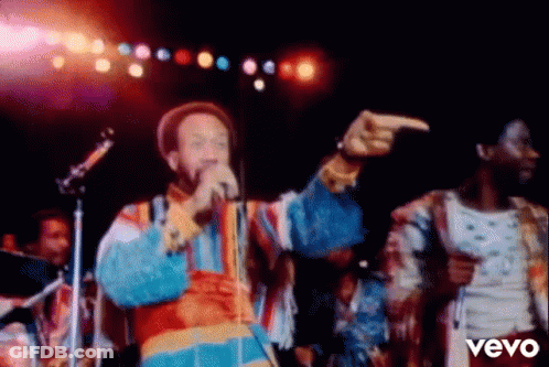 A Man Is Singing Into A Microphone And Pointing At The Camera While A Woman Stands Behind Him . GIF