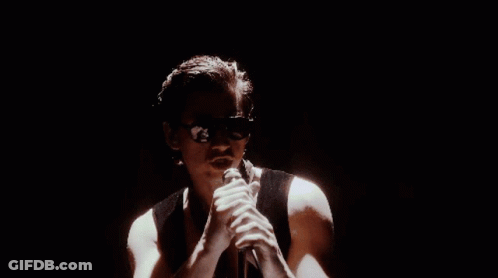 A Man Is Singing Into A Microphone In A Dark Room While Wearing Sunglasses . GIF