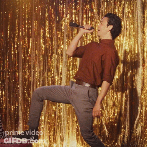 A Man Is Singing Into A Microphone In Front Of A Gold Curtain That Says Prime Video On It GIF