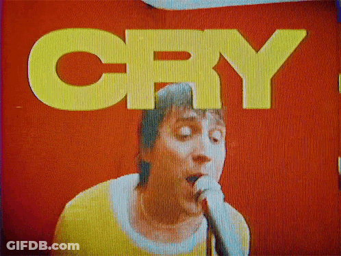A Man Is Singing Into A Microphone In Front Of A Red Wall That Says Cry GIF
