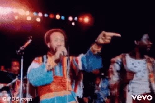 A Man Is Singing Into A Microphone On A Stage While Another Man Points At Him . GIF