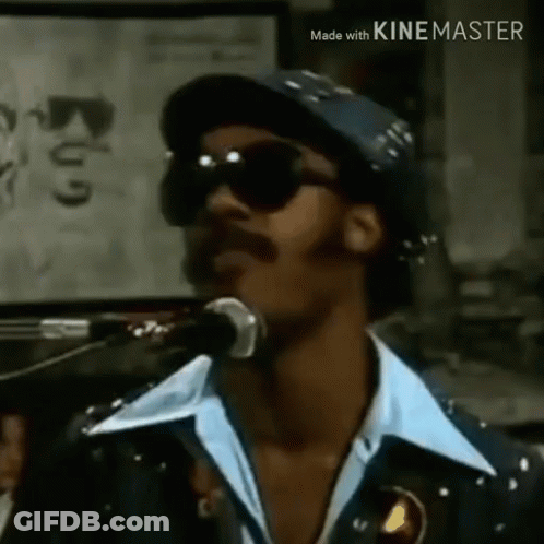 A Man Is Singing Into A Microphone Wearing Sunglasses And A Hat . GIF