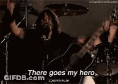 A Man Is Singing Into A Microphone While Holding A Guitar And A Drum Set . GIF