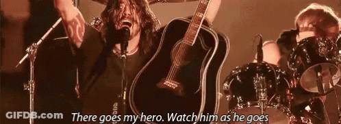 A Man Is Singing Into A Microphone While Holding A Guitar In Front Of A Drum Set . GIF