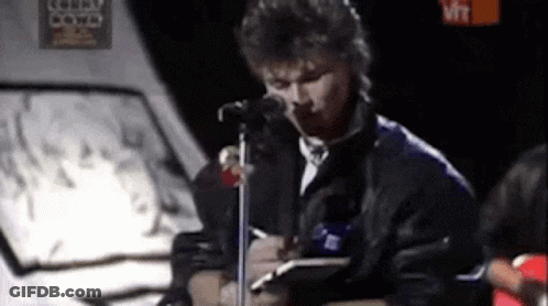 A Man Is Singing Into A Microphone While Holding A Notebook . GIF