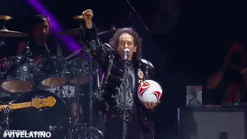A Man Is Singing Into A Microphone While Holding A Soccer Ball On Stage . GIF