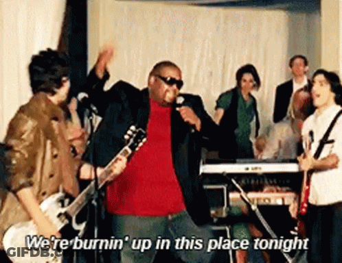 A Man Is Singing Into A Microphone While Playing A Guitar And A Group Of People Are Standing Around Him . GIF