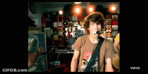 A Man Is Singing Into A Microphone While Playing A Guitar In A Room With Records On The Wall . GIF