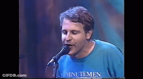 A Man Is Singing Into A Microphone While Wearing A Blue Shirt That Says Minutemen . GIF