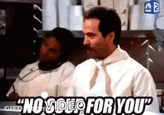 A Man Is Sitting At A Counter In A Kitchen Talking On A Cell Phone While Another Man Looks On . GIF