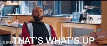 A Man Is Sitting At A Desk In A Kitchen With The Words `` That 'S What 'S Up '' Written Above Him . GIF