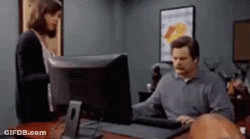 A Man Is Sitting At A Desk In Front Of A Computer While A Woman Stands Behind Him . GIF