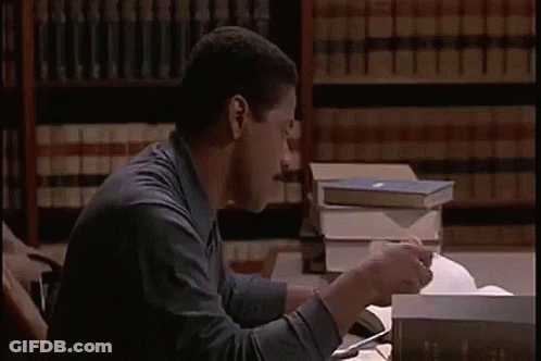 A Man Is Sitting At A Desk Reading A Book And Eating A Sandwich In A Library . GIF