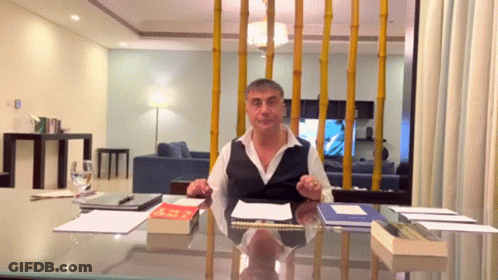 A Man Is Sitting At A Desk With A Book On It And A Glass Of Water In Front Of Him GIF