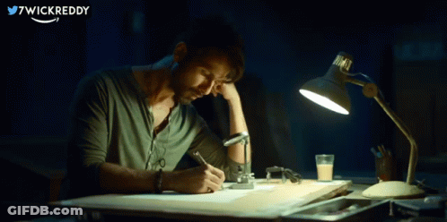 A Man Is Sitting At A Desk With A Lamp And Writing On A Piece Of Paper With 7Wickreddy Written In The Corner GIF