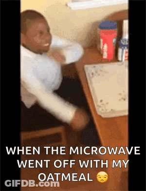 A Man Is Sitting At A Desk With His Arms Outstretched While A Microwave Went Off With His Oatmeal GIF
