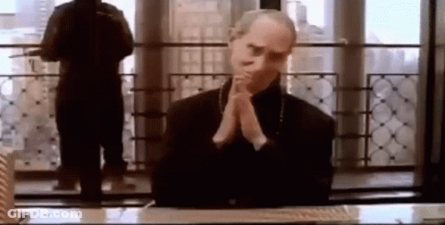 A Man Is Sitting At A Desk With His Hands Folded In Front Of A Window . GIF