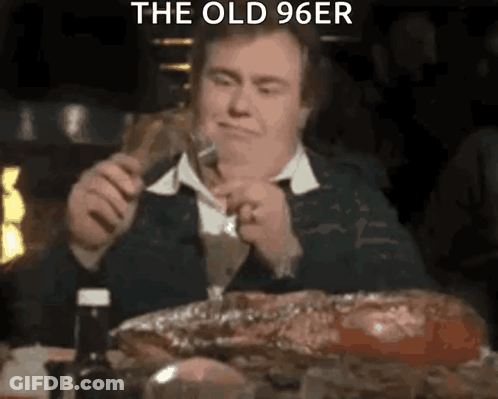 A Man Is Sitting At A Table Eating A Large Piece Of Meat While Holding A Fork . GIF