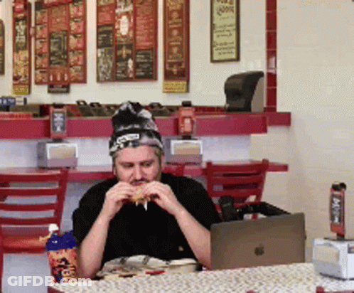A Man Is Sitting At A Table Eating A Sandwich And Drinking A Cup Of Pepsi GIF