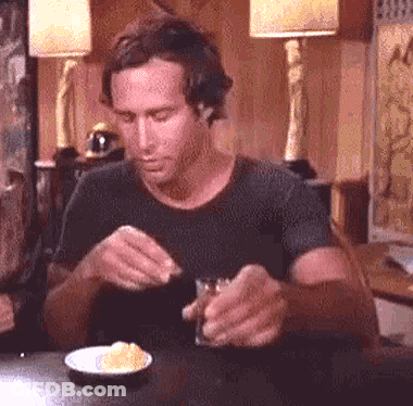 A Man Is Sitting At A Table Eating A Sandwich And Drinking A Glass Of Whiskey . GIF