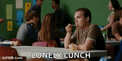 A Man Is Sitting At A Table Eating A Spoonful Of Food In A Cafeteria . GIF